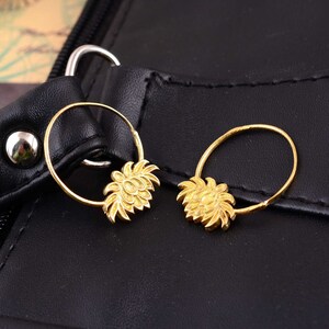 Könnte beinhalten: Ein Paar goldene Creolen mit einem floralen Design. Die Ohrringe liegen auf einer schwarzen Lederoberfläche.