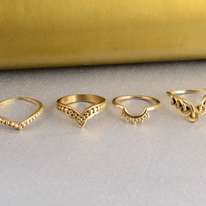 Könnte beinhalten: Vier goldfarbene Ringe mit einzigartigen Designs. Die Ringe variieren in der Form, darunter eine V-Form, ein Halbkreis mit kleinen Perlen und ein offenes Design mit dekorativen Elementen. Die Ringe werden auf einer hellgrauen Oberfläche präsentiert.