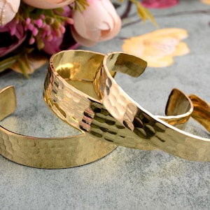 Könnte beinhalten: Drei goldfarbene, gehämmerte Metallarmbänder, die übereinander gestapelt sind. Die Armbänder haben alle die gleiche Breite und eine leicht abgerundete Form.