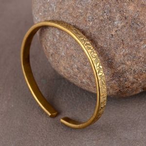 May include: A gold-toned metal bangle bracelet with an intricate engraved pattern. The bracelet is open and has a simple, minimalist design.