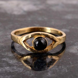 Könnte beinhalten: Ein goldfarbener Ring mit einem augenförmigen Design. Der Ring hat einen schwarzen, runden Edelstein in der Mitte des Auges. Der Ring wird auf einer reflektierenden Oberfläche gezeigt.
