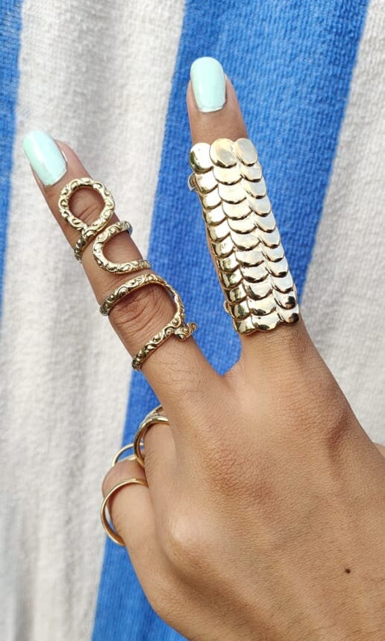 May include: A set of gold-toned rings, including a snake-shaped ring and a ring with a chainmail design. The rings are worn on a hand with light blue nail polish.