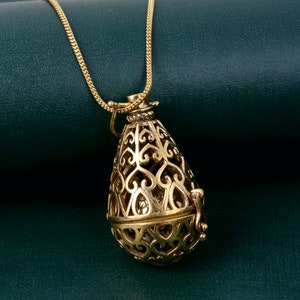 Könnte beinhalten: Eine goldfarbene Halskette mit einer zarten Kette und einem Anhänger in Form eines verzierten Eies. Der Anhänger ist durchbrochen gearbeitet und zeigt ein detailliertes Design aus Herzen und Spiralen.