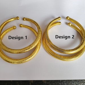 Könnte beinhalten: Zwei goldfarbene Armreifen, einer mit einer strukturierten Oberfläche und der andere mit einem filigranen Blumenmuster. Die Armreifen sind mit "Design 1" und "Design 2" beschriftet.