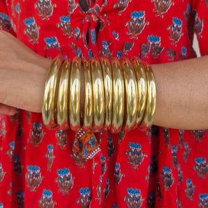 以下が含まれることがあります： 10個の金色の金属製のバングルが積み重ねられています。バングルは人の手首に重ねられています。