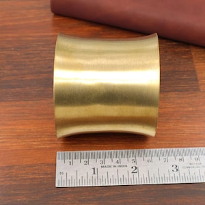 Könnte beinhalten: Ein breites, gebogenes Armband aus Messing. Das Armband hat eine glatte, goldene Farbe. Ein Metalllineal mit Zollmaßen liegt neben dem Armband. Der Text "MADE IN INDIA" ist auf dem Lineal zu sehen.