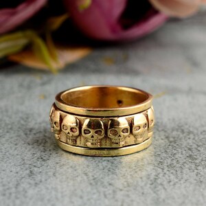 Könnte beinhalten: Ein goldfarbener Ring mit einer Reihe von Totenköpfen um den Umfang. Die Totenköpfe sind detailliert und haben ein leicht erhabenes Design.
