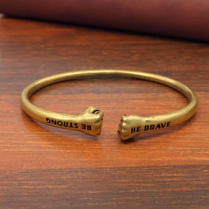 Könnte beinhalten: Ein goldfarbenes Armband mit den Worten "BE STRONG" und "BE BRAVE" an den Enden. Das Armband hat Faust-förmige Enden und wird auf einer Holzoberfläche präsentiert.