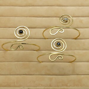 May include: Three gold-toned arm cuffs with a spiral design and gemstone accents. The cuffs feature a curved, open design with a central spiral and a gemstone. The gemstones are black and light gray.