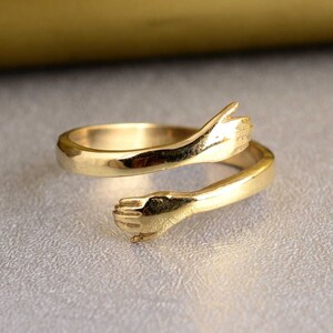 Könnte beinhalten: Ein goldener Ring mit zwei ineinander verschlungenen Händen, die einen Kreis bilden. Der Ring ist aus glattem, glänzendem Metall gefertigt und hat ein schlichtes, elegantes Design.
