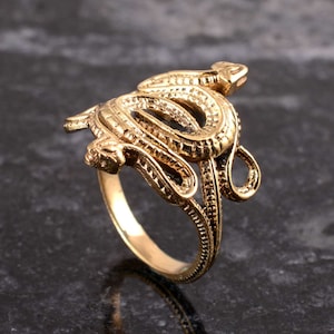 Könnte beinhalten: Ein goldfarbener Ring mit einem detaillierten Schlangendesign. Die Schlange ist zusammengerollt und ihr Kopf ist erhoben, was einen dreidimensionalen Effekt erzeugt.