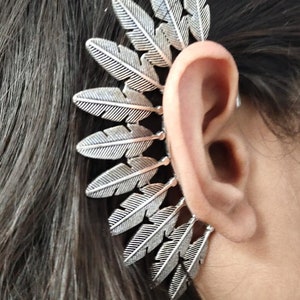 Könnte beinhalten: Silberner Feder-Ohrschmuck. Der Ohrschmuck besteht aus mehreren Federn, die in Fächerform angeordnet sind. Der Ohrschmuck wird am oberen Teil des Ohrs getragen.
