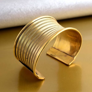 Könnte beinhalten: Ein goldfarbenes Metall-Armband mit geflochtenem Design. Das Armband ist offen und hat eine leicht gebogene Form.