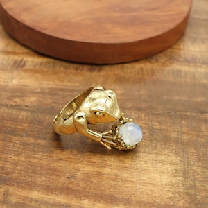 Könnte beinhalten: Ein goldfarbener Froschring, der einen runden, hellblauen Edelstein hält. Der Ring ist mit strukturierten Oberflächen und einem skurrilen Design versehen. Die Arme des Frosches umfassen den Edelstein.