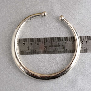 Könnte beinhalten: Ein silbernes Armreif-Armband mit abgerundeten Kanten, das einen Durchmesser von etwa 17,8 cm hat. Das Armband wird neben einem Lineal zur Größenreferenz gezeigt.