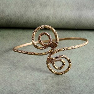 Könnte beinhalten: Ein bronzefarbenes Armband mit Schlangenmuster. Das Armband hat zwei spiralförmige Schlangenköpfe und ein Band mit sich wiederholendem Blättermuster. Das Armband wird vor einem gedämpften grünen Hintergrund gezeigt.