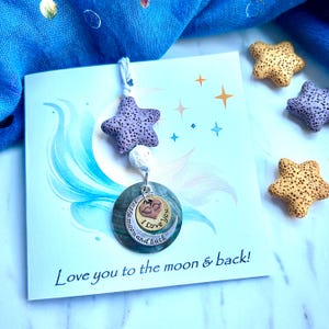 May include: A purple star charm with a white bead and a silver charm with a crescent moon and a heart with wings. The charm says "to the moon and back I love you". The charm is on a blue card with white stars and the text "Love you to the moon & back!"
