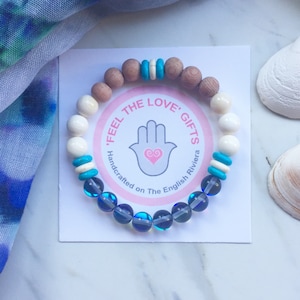 May include: A blue and white beaded bracelet with a pink and white tag that says "Feel the Love' Gifts Handcrafted on The English Riviera".