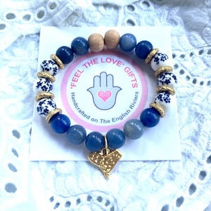 May include: Blue and white ceramic beads with gold accents, blue agate beads, and a wooden bead on a bracelet with a gold charm in the shape of a heart. The bracelet has the text "Feel the Love Gifts Handcrafted on The English Riviera" on a pink background.