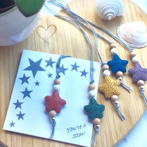 May include: Five colourful star-shaped lava stone necklaces with wooden beads and grey cords. The necklaces are arranged on a wooden surface with a card that says "You're a Star!"