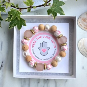 May include: A pink and white floral bracelet with wooden beads, gold accents, and a pearl charm. The bracelet is in a white box with a card that reads "Feel the Love Gifts Handcrafted on The English Riviera".