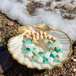 May include: Two bracelets on a white seashell with a gold rim. One bracelet is made of light brown wooden beads, and the other is made of green and white beads.