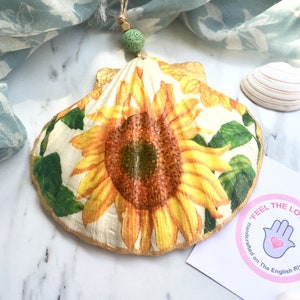 May include: A painted seashell decoration with a sunflower design. The sunflower has yellow petals and a brown centre, with green leaves. It hangs from a jute string and a green bead. A small tag with text and a logo is visible.