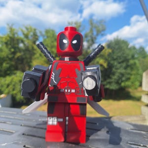 May include: A red and black plastic toy figure resembling a comic book character with a mask, white eye patches, and a torso printed with a belt and chest details. The figure holds two black blocky toy guns and has two sword hilts visible on its back.