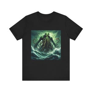Puede incluir: Camiseta negra con un diseño de una criatura verde con tentáculos emergiendo de las olas del océano bajo un cielo tormentoso. El diseño muestra una figura humanoide con cabeza de pulpo. La obra de arte está centrada en la camiseta.