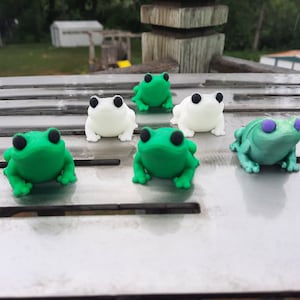 May include: Six small frog figurines, three green and three white, with black eyes. The frogs are arranged on a silver surface.