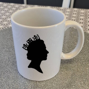 Könnte beinhalten: Eine weiße Keramiktasse mit einer schwarzen Silhouette einer Person mit einer Krone.