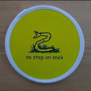 Pode incluir: Um remendo circular amarelo com uma borda branca. O remendo apresenta uma cobra preta e verde com o texto "no step on snek".