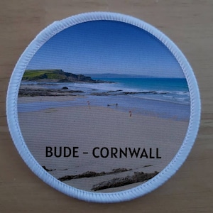 May include: A white round patch with a photo of a beach scene with blue water and a sandy beach. The text "BUDE - CORNWALL" is printed in black on the patch.