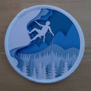 Może przedstawiać: Biała haftowana naszywka z niebieskim tłem przedstawiająca sylwetkę wspinacza wspinającego się po skalnej ścianie z liną. Wspinacz jest sylwetką na tle niebieskiego nieba z górami w tle. Dolna część naszywki przedstawia las drzew.