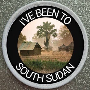 May include: A white round patch with a black circle in the center. The black circle has the text "I've Been To South Sudan" in white text. The center of the circle shows a photo of a village in South Sudan.