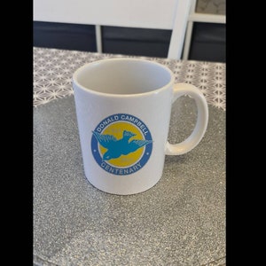 Könnte beinhalten: Weiße Keramiktasse mit einem blauen und gelben Logo, auf dem "Donald Campbell Centenary" steht und ein blauer Vogel im Flug zu sehen ist.