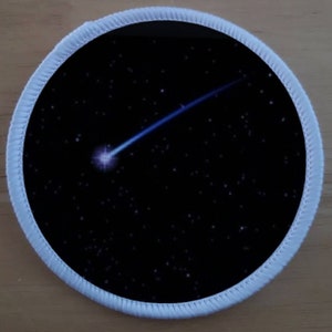 Puede incluir: Un parche redondo blanco con fondo negro que presenta una estrella fugaz blanca con una cola azul en un cielo estrellado azul oscuro.