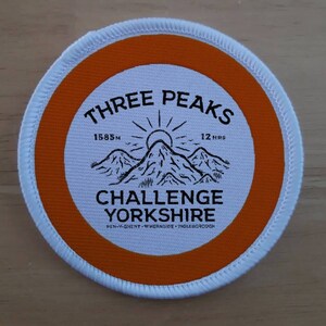 Op de afbeelding: Een ronde witte en oranje patch met de tekst "THREE PEAKS" in zwarte letters. De patch bevat ook een zwart-witte illustratie van drie bergen met een zon erboven. De tekst "1585m" en "12 HRS" zijn ook in zwarte letters opgenomen. De tekst "CHALLENGE YORKSHIRE" is in zwarte letters. De tekst "PEN-Y-GHENT-WHERNSIDE-INGLEBOROUGH" is in zwarte letters.
