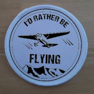 Puede incluir: Parche redondo blanco con texto negro y una silueta negra de un avión pequeño. El texto dice "I'd Rather Be Flying".
