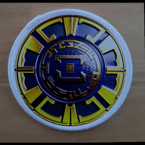 May include: A white circular patch with a gold and blue emblem. The emblem features a stylized letter "E" surrounded by a gold design. The patch has a white border with a gold stitching.