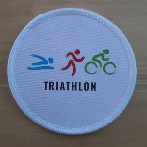 Puede incluir: Un parche redondo blanco con la palabra "TRIATHLON" impresa en negro. El parche presenta tres iconos que representan los tres deportes de un triatlón: natación, carrera y ciclismo. Los iconos son de color azul, rojo y verde.