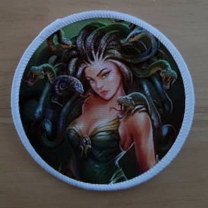 May include: A round, white patch with a colorful image of Medusa, a mythical Greek figure with snakes for hair, surrounded by green snakes.