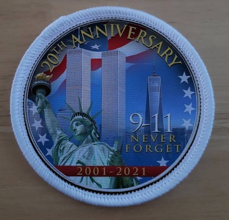 May include: A white round patch with a blue, red, and white image of the Twin Towers and the Statue of Liberty. The text "20th Anniversary 9-11 Never Forget 2001-2021" is printed on the patch.