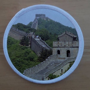 May include: A round patch with a white border featuring a photograph of the Great Wall of China. The wall is made of stone and winds its way up a hill. There are watchtowers along the wall, and some people are walking on the wall.