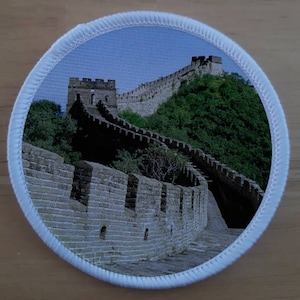 May include: A round patch with a white border and a printed image of the Great Wall of China. The wall is made of stone and is winding up a hill. The sky is blue and there are trees in the background.