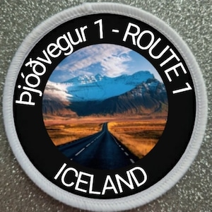 May include: A white circular patch with a black circle in the center. The black circle has a picture of a road leading through a mountain landscape with a blue sky and white clouds. The text "Ring Road 1 - Route 1" is written in white around the black circle. The text "Iceland" is written in white at the bottom of the black circle.
