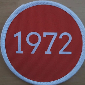 Może przedstawiać: Okrągła naszywka z czerwonym środkiem i białym obramowaniem. Numer "1972" jest wydrukowany na biało, wyśrodkowany na czerwonym tle. Naszywka ma teksturowany wygląd.