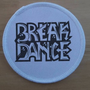 May include: White round fabric patch with black text that reads "Break Dance" in a graffiti style font.