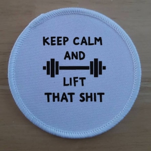 Könnte beinhalten: Runder weißer Patch mit schwarzem Text, der "Keep Calm and Lift That Shit" lautet, mit einer schwarzen Hantelgrafik.