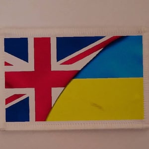 Può includere: Una bandiera rettangolare con un design che combina la Union Jack del Regno Unito con il blu e il giallo della bandiera ucraina. La bandiera ha un bordo bianco con un bordo cucito.